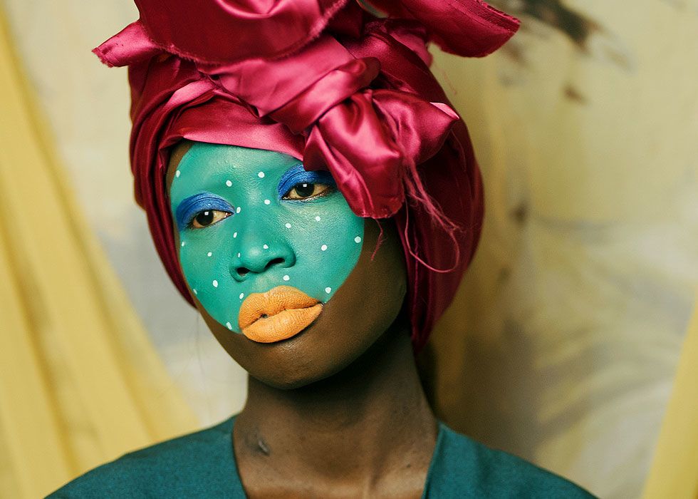 Close up of a dark skinned woman looking away from the camera. She is wearing a maroon coloured head scarf and her face is painted teal with white dots, her lips are painted peach and her eyelids are painted in blue