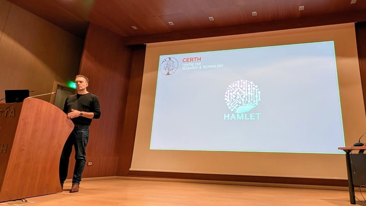 A man standing beside a podium and in front of projector screen with a logo "HAMLET" displayed