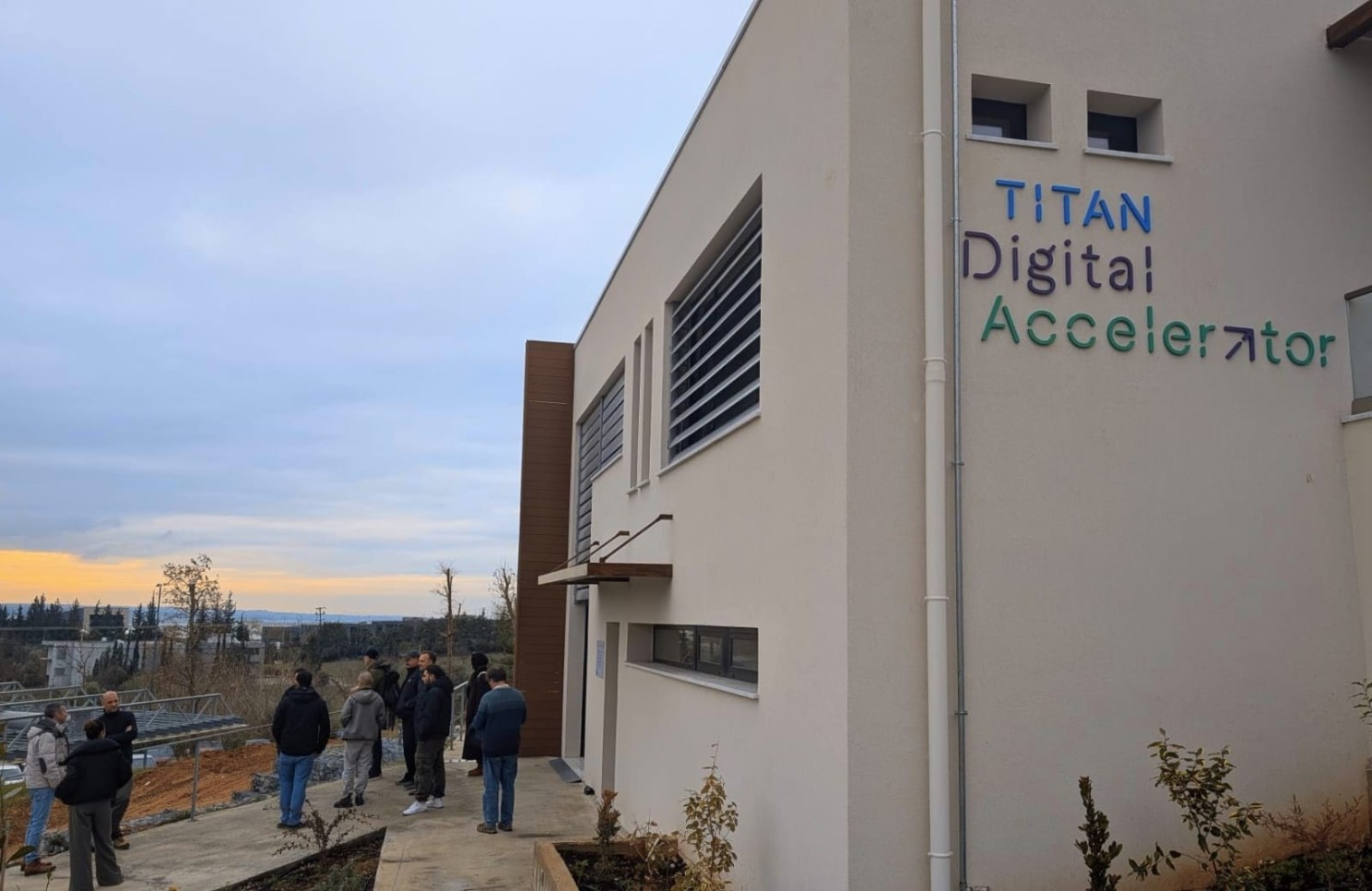 A group of people stands outside a modern building labelled "Titan Digital Accelerator" with a cloudy sky and distant trees in the background.