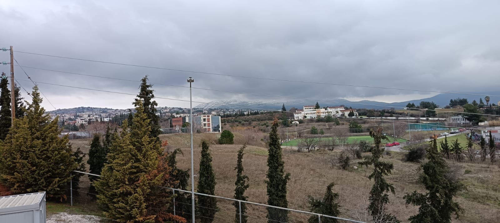 An image of rural landscape with scattered buildings, trees, and a fenced area. Hills and a small town are visible in the background.