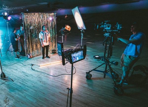 Image of a man standing in front of cameras with string light background. 