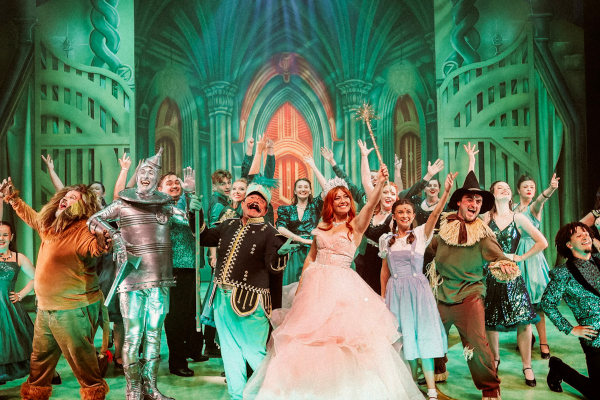 Wizard of Oz performers on stage with their arms stretched upwards in front of a green background