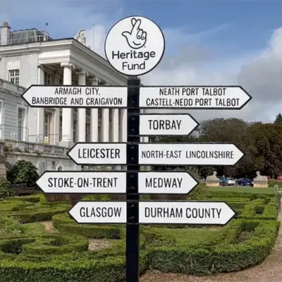 Sign post with "Heritage Fund" at the top, showing signs to 9 places: Stoke on Trent (Midlands & East) Leicester (Midlands & East) County Durham (North) North-East Lincolnshire (North) Torbay (London & South) Medway (London & South) Glasgow (Scotland) Armagh City, Banbridge and Craigavon (Northern Ireland) Neath Port Talbot (Wales)
