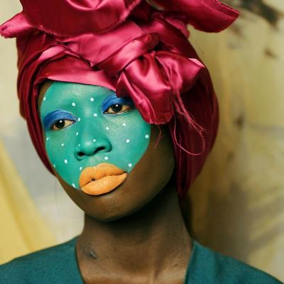 Close up of a dark skinned woman looking away from the camera. She is wearing a maroon coloured head scarf and her face is painted teal with white dots, her lips are painted peach and her eyelids are painted in blue