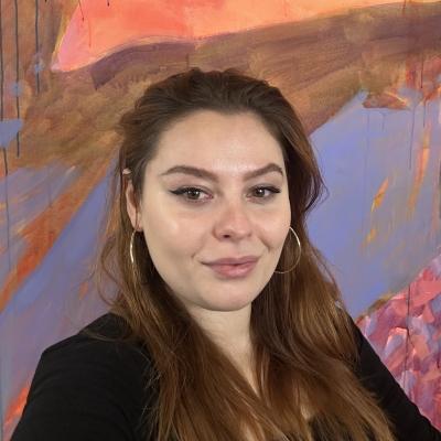A portrait photograph of a white woman with long brown hair wearing earrings in front of a colourful background