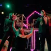 Image of 3 artists performing on a dimly lit stage with pink neon lights. One man is singing and one female appears to be dancing arms raised 